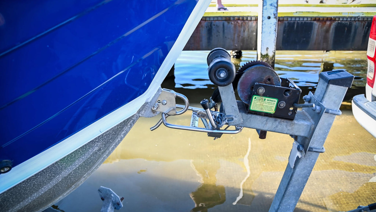 Close-up of the EasiLaunch Boat Catch system installed on a metal boat trailer's winch mount, latched securely to a blue boat's bow. Outdoor waterfront and dock structures are visible in the background.