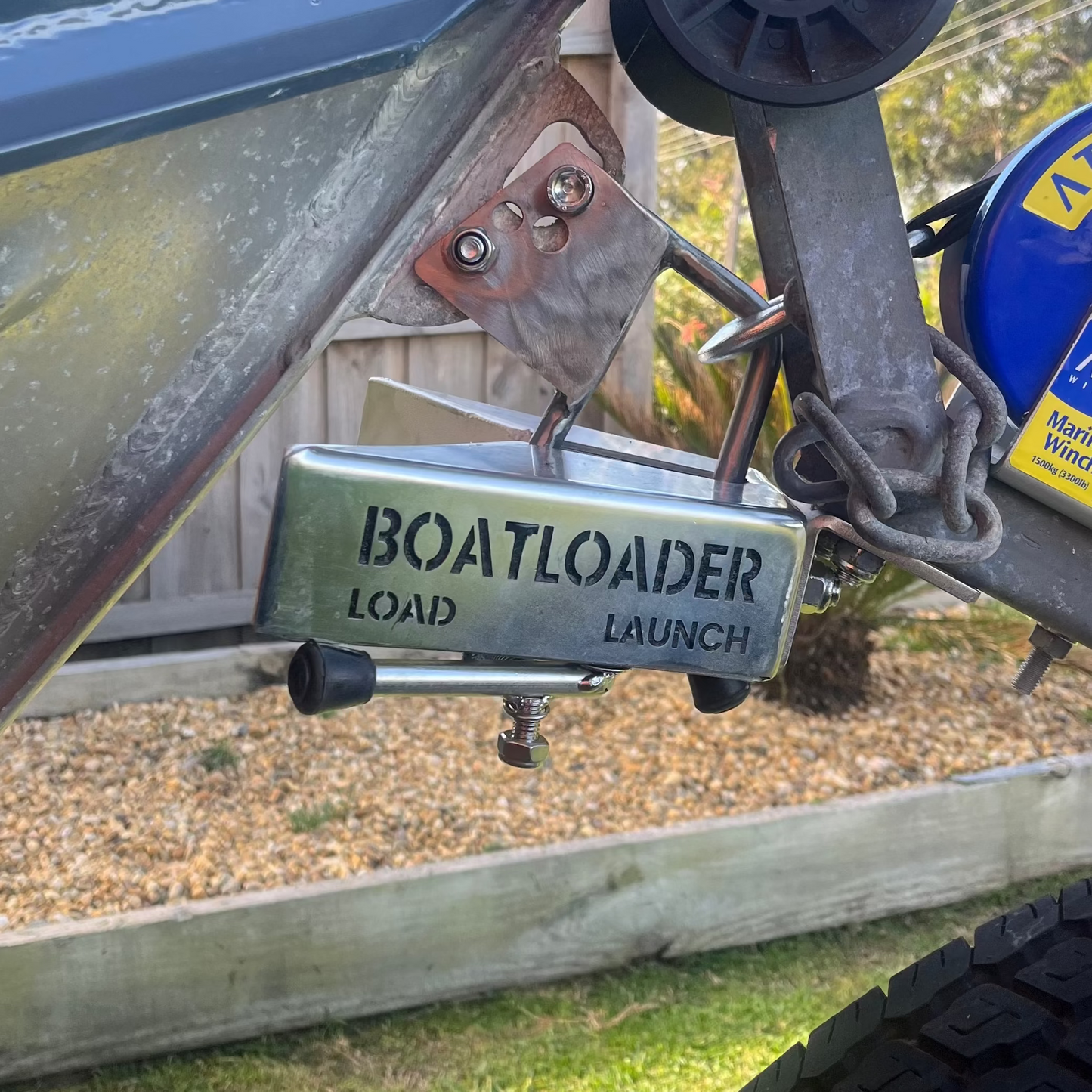 Close-up of a boat trailer hitching to a launch or boat, with a metal plate reading 'BOATLOADER LOAD LAUNCH' and part of an Atlantic trailer wheel visible outdoors on grass.
