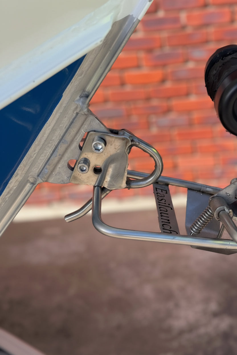 Close-up of the EasiLaunch Boat Catch system installed on a metal boat trailer's winch mount, latched securely to a blue and white boat's bow. Outdoor red brick wall and concrete driveway are visible in the background.